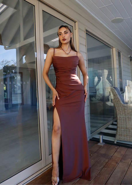 A woman models a floor-length, rust-brown strapless gown with a high thigh slit, standing on a wooden deck in front of glass patio doors.