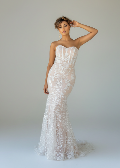 A woman modeling a strapless mermaid-style wedding gown with a sweetheart neckline, fitted lace bodice, and flared lace skirt on a light gray studio background.